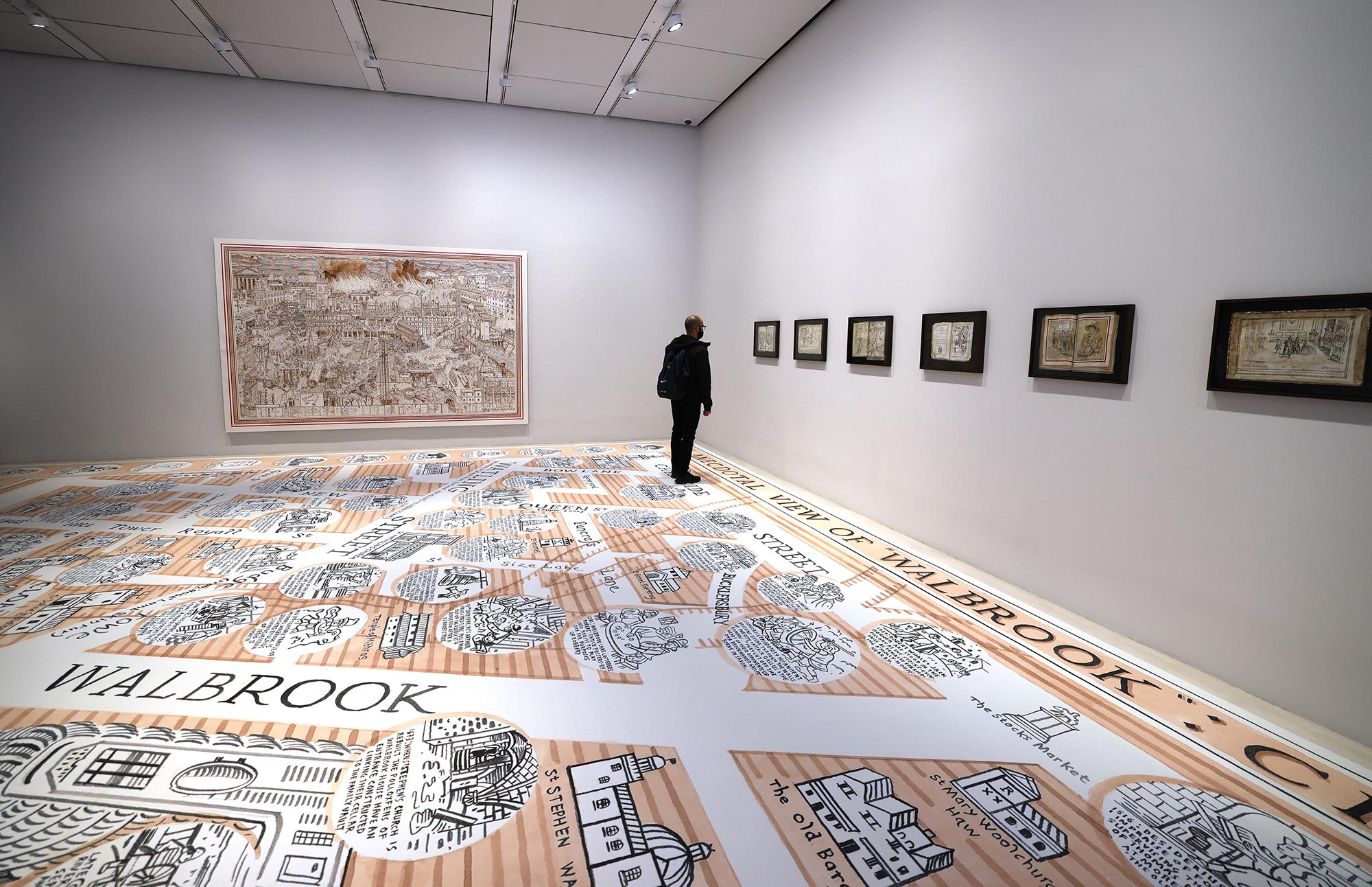 An guest of Bloomberg SPACE at London Mithraeum looks at a smaller Adam Dant art piece, a part of a row of smaller art pieces at the exhibit filling a wall.