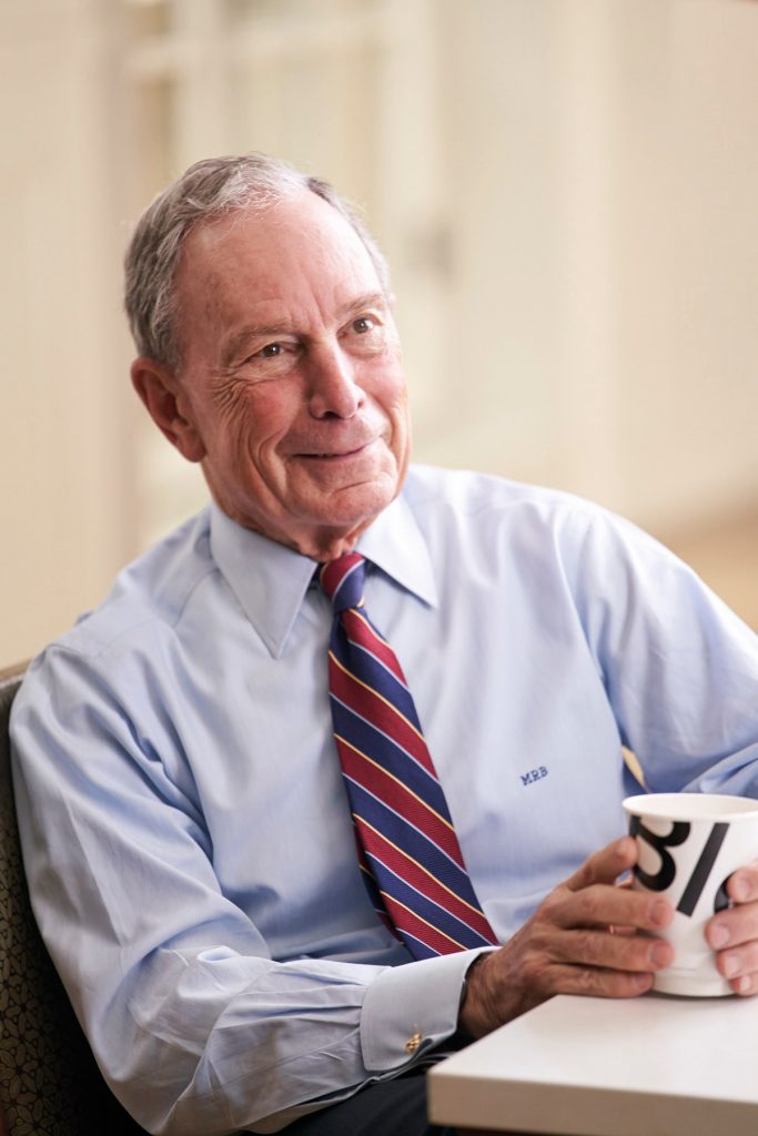Portrait of Mike Bloomberg sitting and holding a coffee cup that says Bloomberg