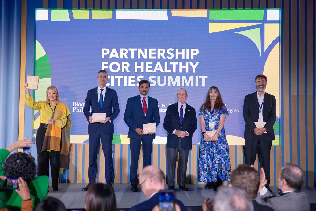 Two women and four men including Mike Bloomberg standing on stage in front of a sign that says "Partnership for Healthy Cities Summit", they are holding boxes with a ribbon.