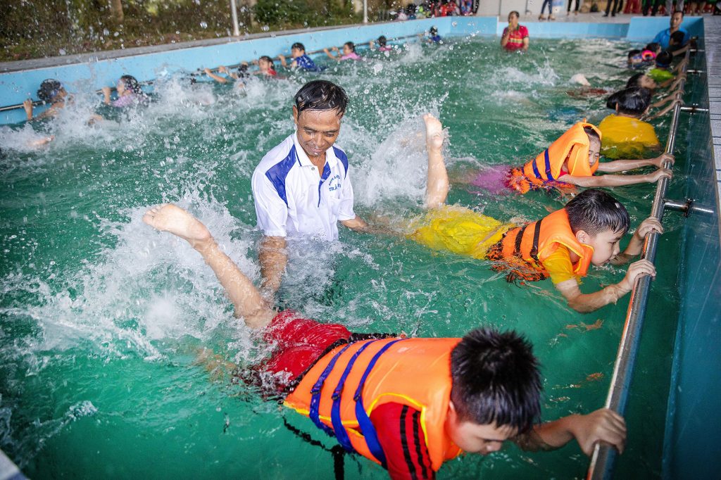 A swimming pool full of kids wearing inflatable orange vests and holding the guard rail while swimming, a male teacher is in the middle of the pool.