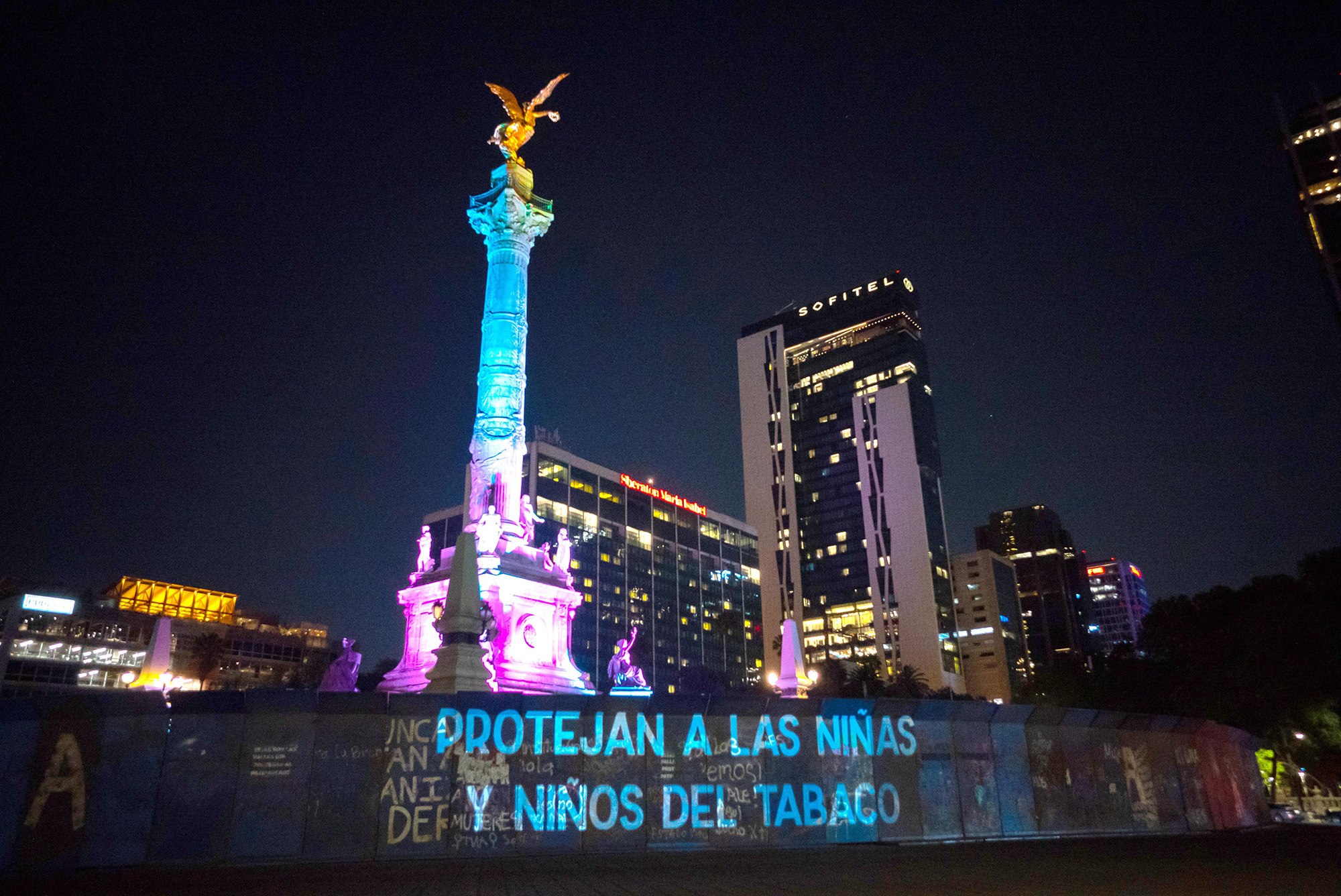 Photo of a column lit up with green, blue and purple light at night with sky scrapers behind. A fence in front of the columns says "Protect our girls and boys from tobacco" in Spanish.