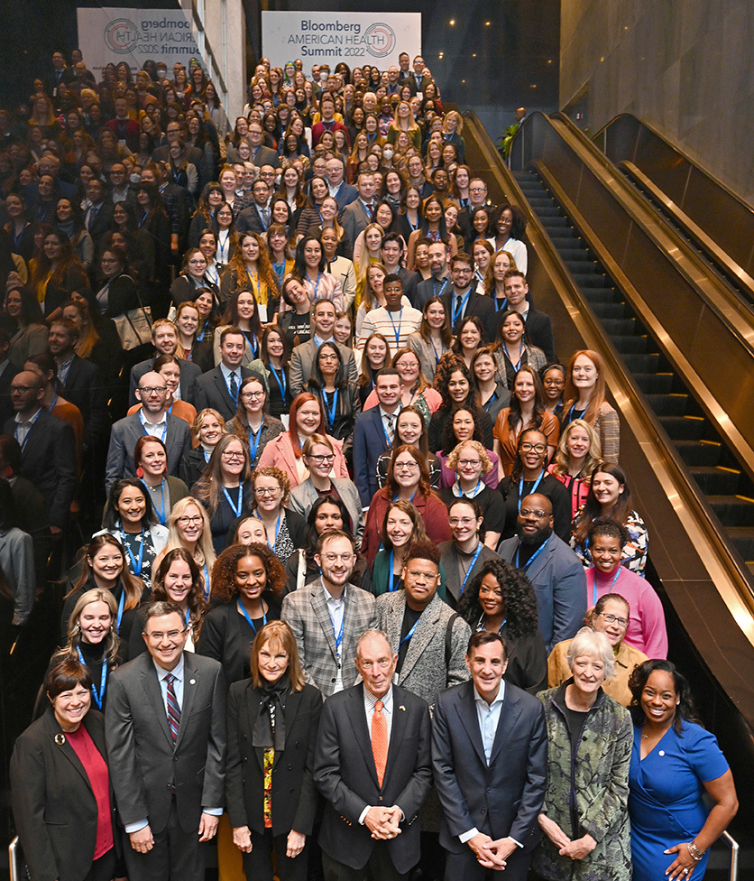 Group photo of a diverse group of people standing on a long staircase inside a building. A poster behind them says "Bloomberg American Health Summit 2022".