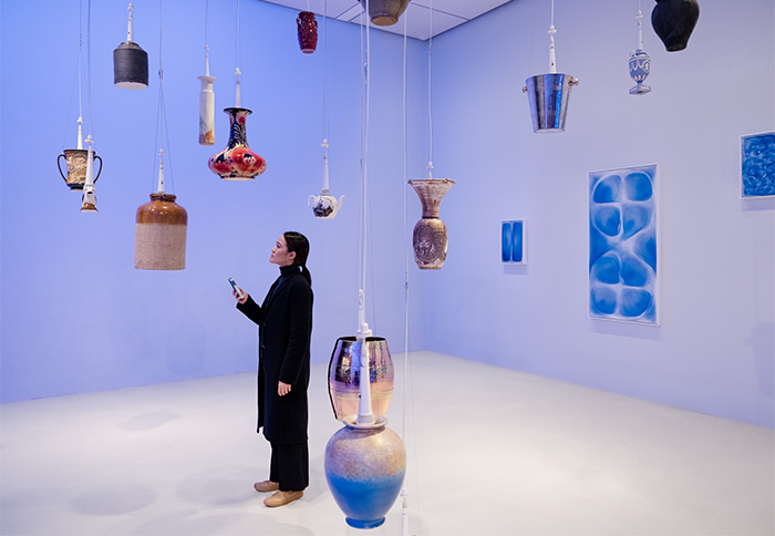 Photo of a woman in a white room with vases and vessels hanging from the ceiling, woman is holding her phone out
