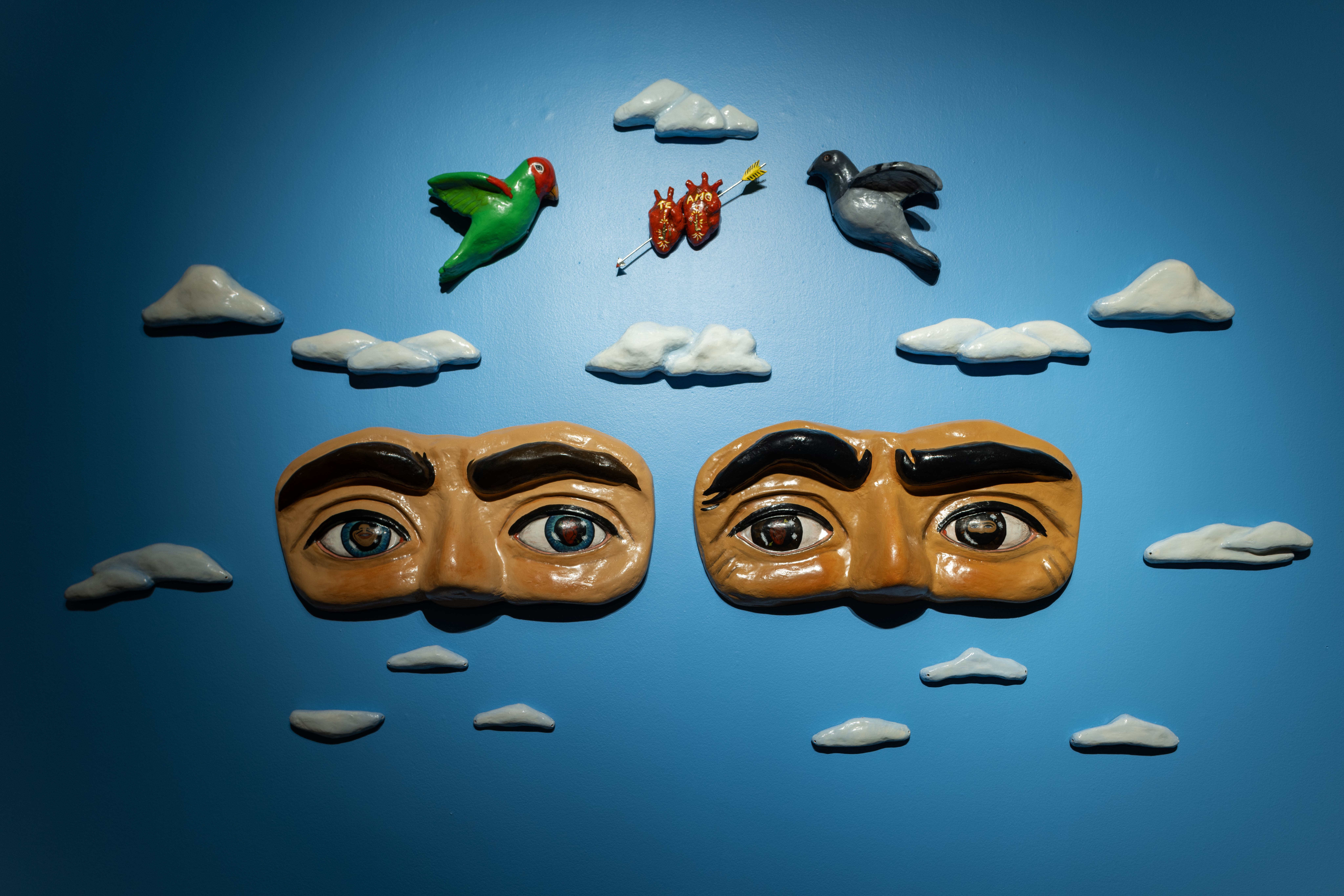 Photo of Joel Hernandez’s At First Sight. Two paper mache sculpture of faces set on a bright blue background surrounded by paper mache clouds, birds and hearts.