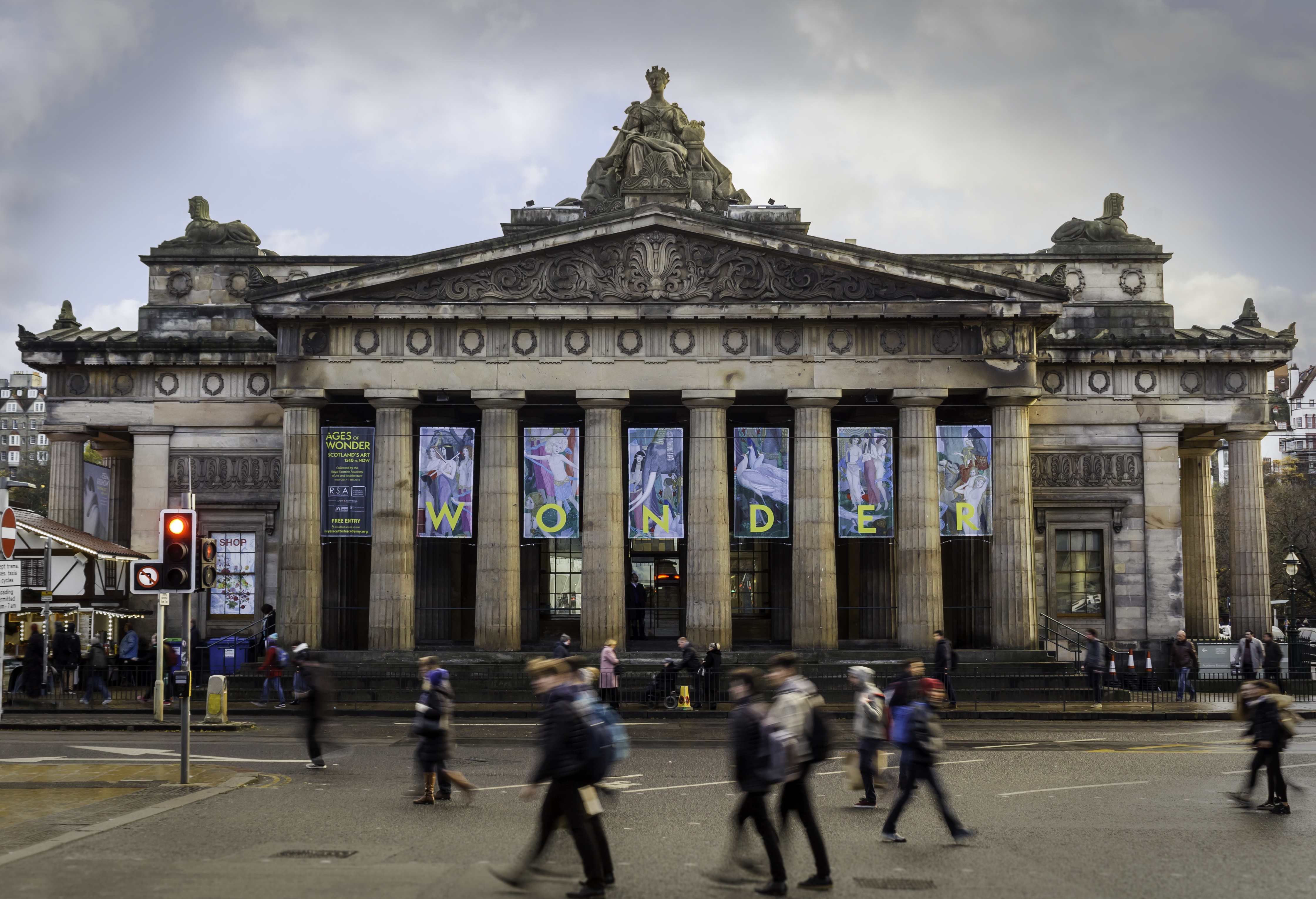 Exterior shot of Royal Scottish Academy. Column-fronted building with hanging banners advertising Ages of Wonder Exhibition. People crossing the round out front.