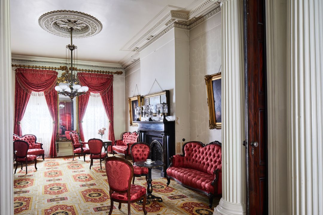 The front parlor contains groups of chairs and couches of matching red satin upholstery. The gasolier and open double windows illuminate the parlor. Gilt mirrors and frames hang on the walls.