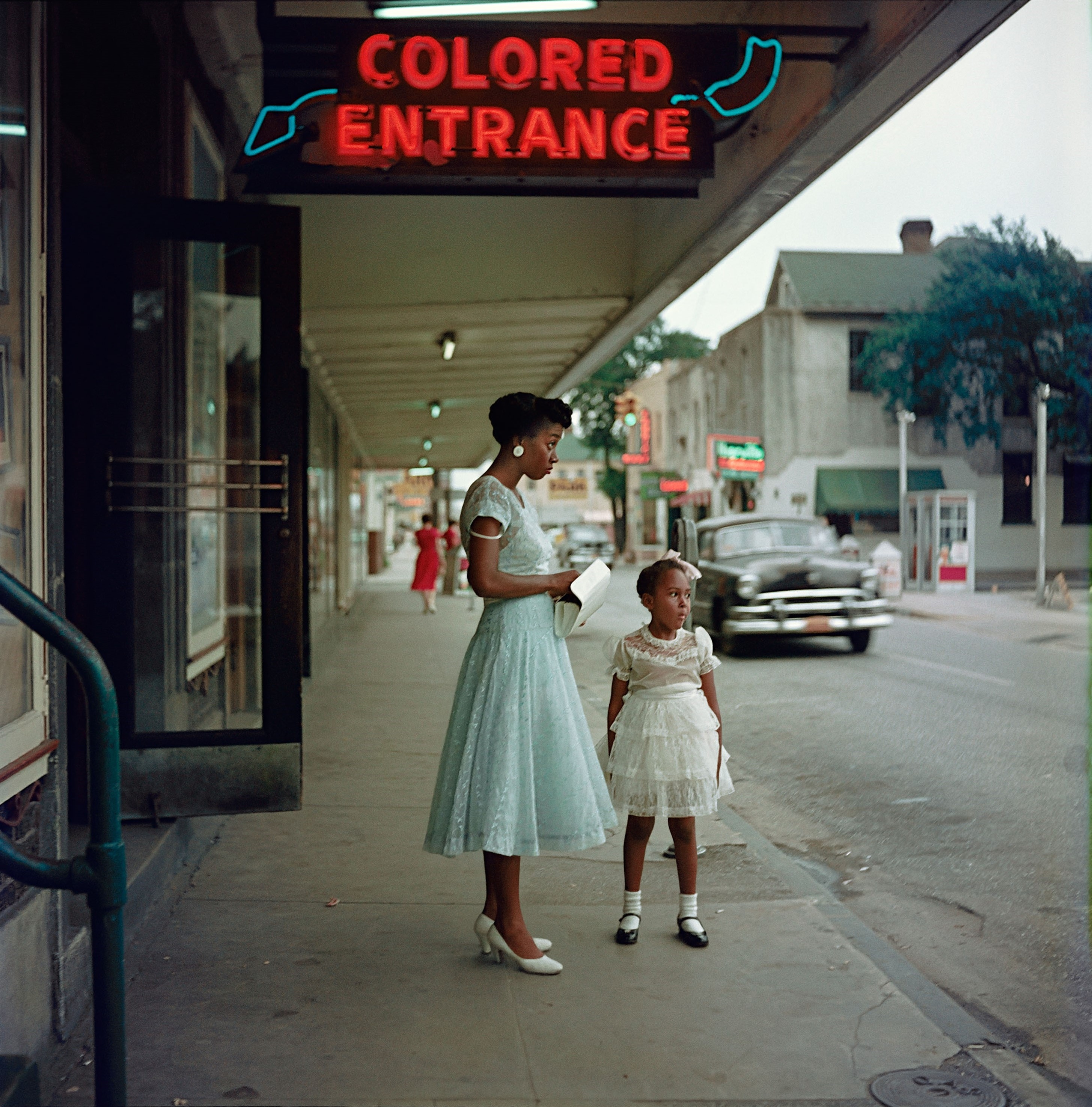 50s color photo with a black woman and her young niece dressed in frilly dresses, standing in front of a theater underneath a sign that reads “colored entrance”.