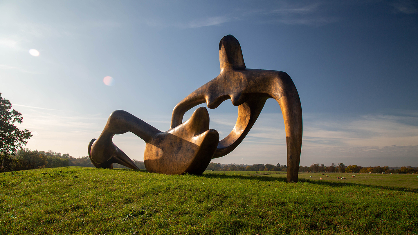 A monumental bronze sculpture of an abstract reclining figure, shown in a landscape setting, resting on grassland, with blue sky behind.