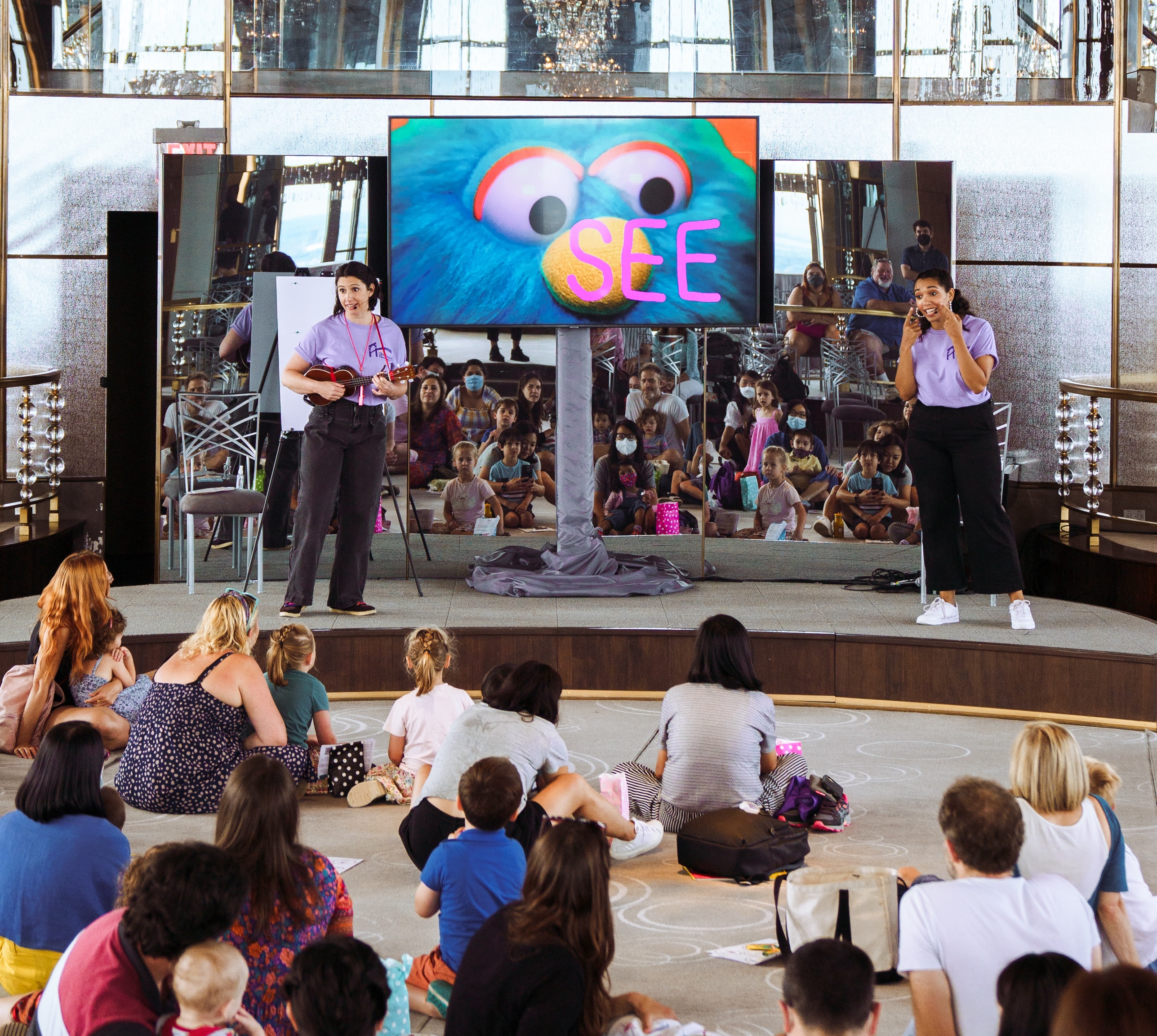 Two women in light purple t-shirts sing and play ukelele on either side of a large video screen which displays a large close up of a bright blue puppet. An audience watches.