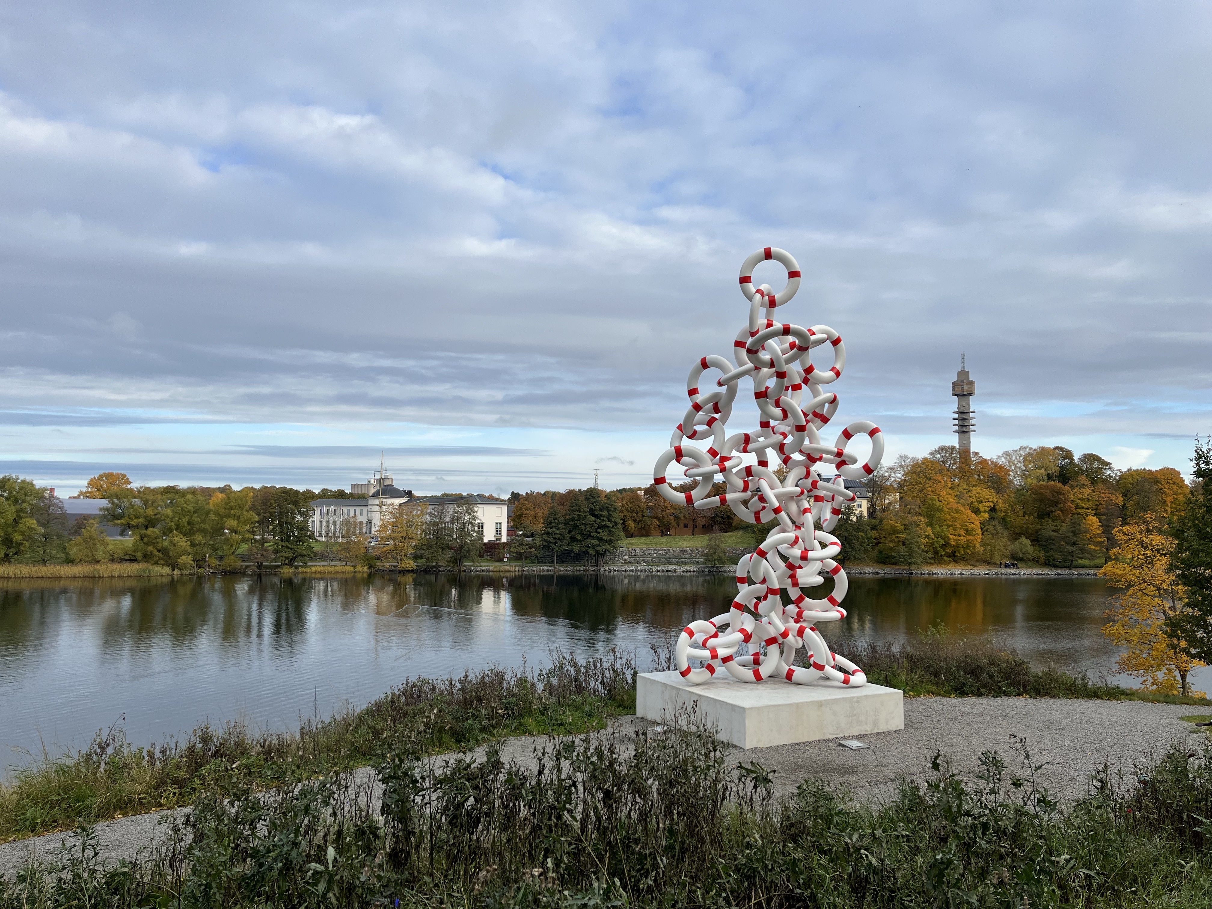 Sculpture of life buoys standing alongside a canal, trees with orange leaves along the shoreline