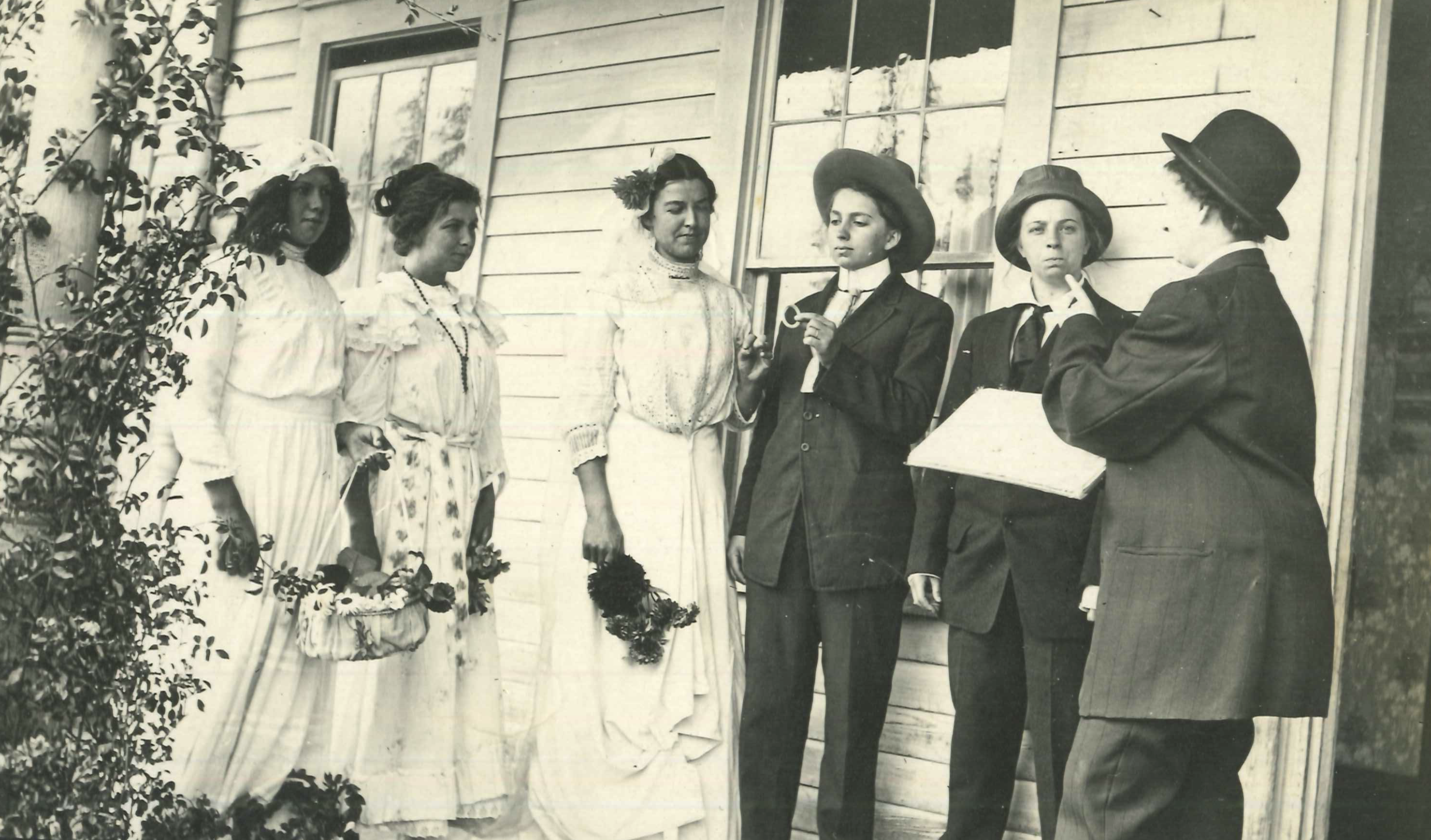A vintage black-and-white photo of a staged wedding scene with three people in bridal gowns and two in suits. One suited individual holds a ring, while an officiant gestures seriously. The group stands on a wooden porch, creating a playful yet formal moment.