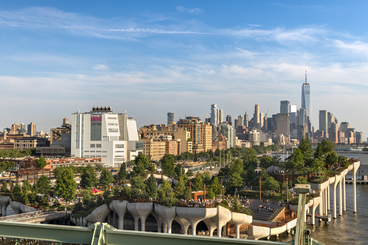 A view of Little Island park on the Hudson River, featuring unique tulip-shaped concrete supports, lush greenery, and people gathered. The New York City skyline and One World Trade Center rise in the background under a blue sky.