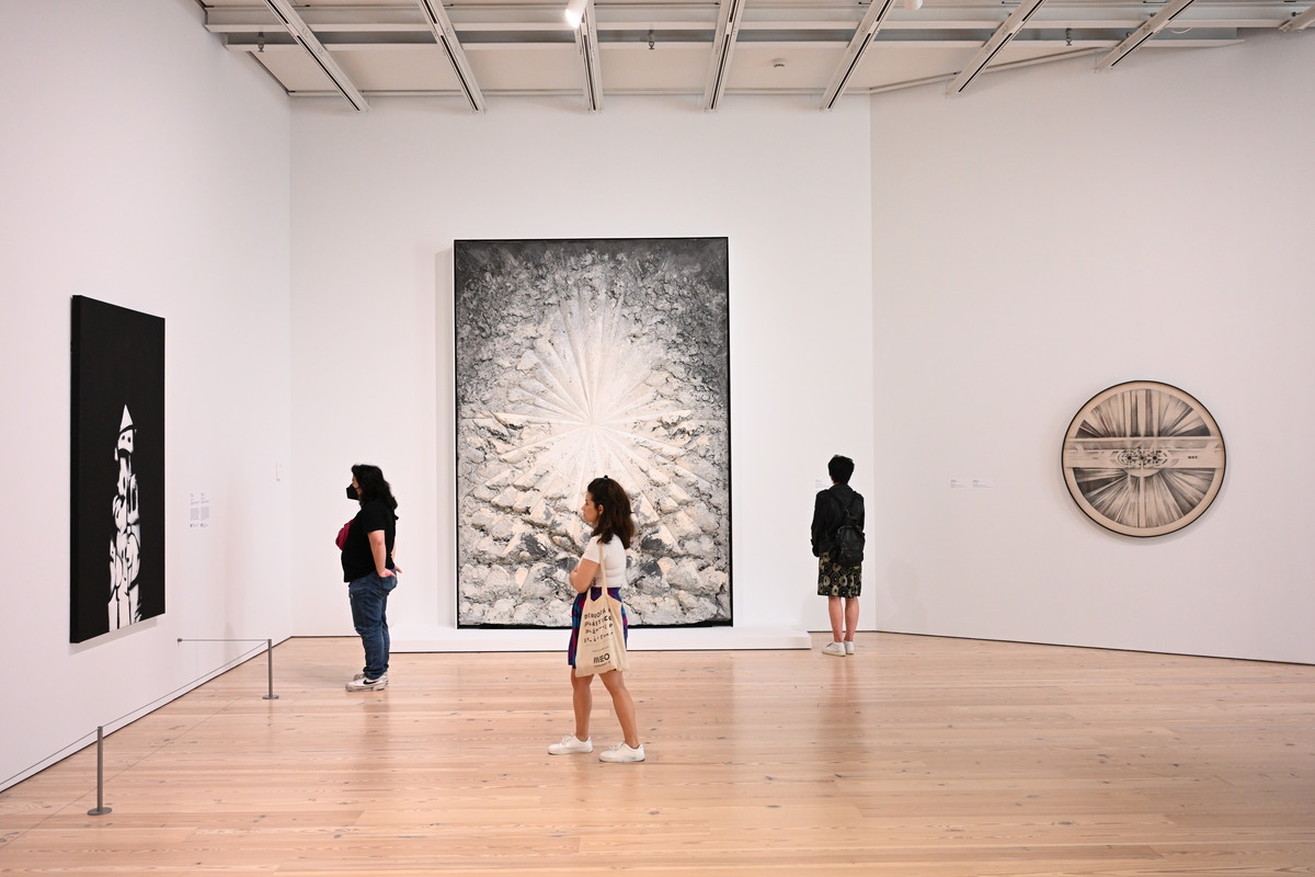 Visitors in a white-walled gallery look at a black painting, a sculpture beige painting, and a circular artwork on the wall.