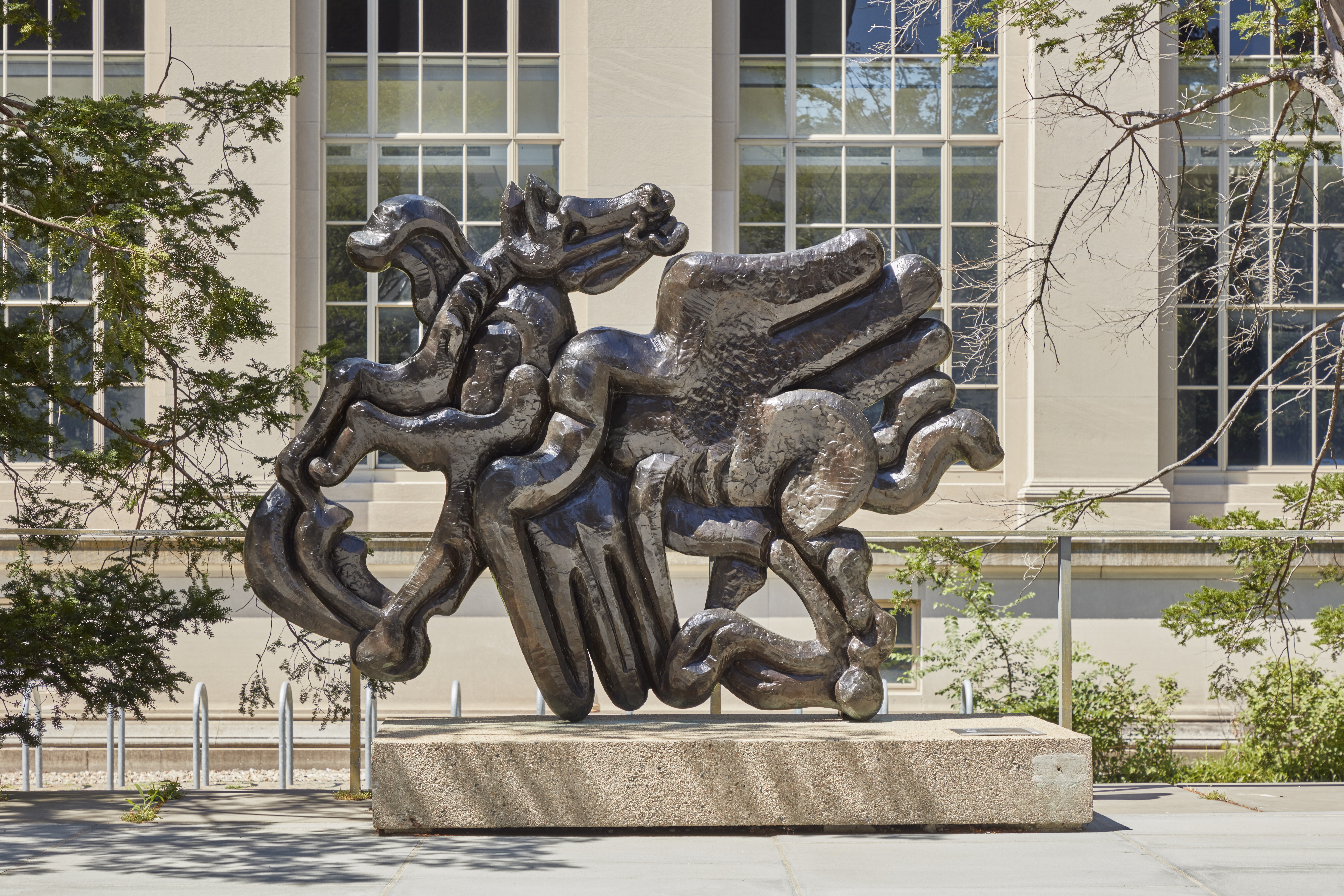 Bronze sculpture on a pedestal featuring a pegasus figure with the horse head facing backwards against a large stone building with long windows.