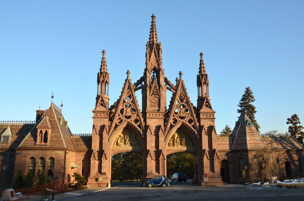A reddish-brown Gothic Revival Arch with multiple spires and carved figures stands under a clear blue sky. Two smaller buildings with patterned roofs flank the ornate entrance.