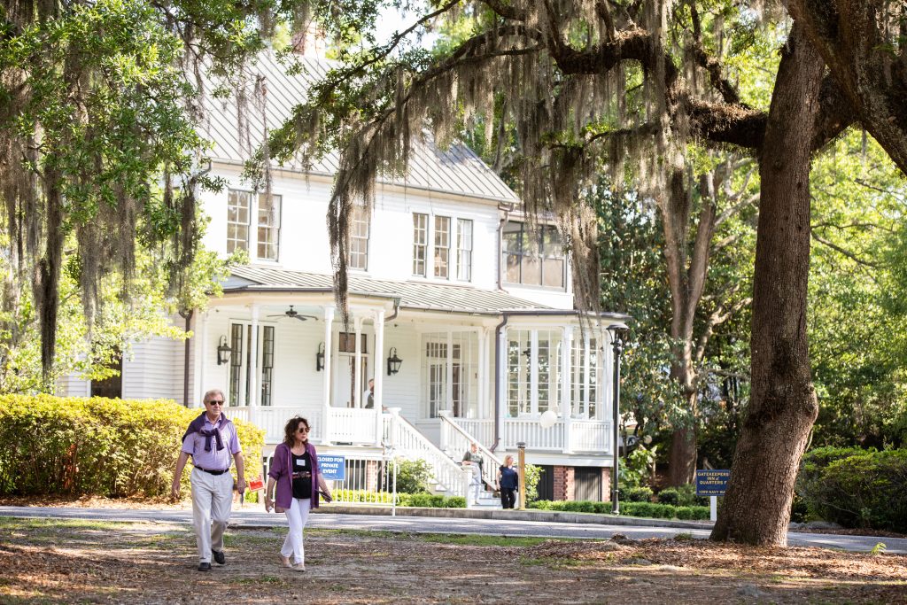 The image shows a large white house with a wraparound porch, partially shaded by large oak trees draped with Spanish moss. Two people are walking on a sidewalk in front of the house, with sunlight filtering through the trees.