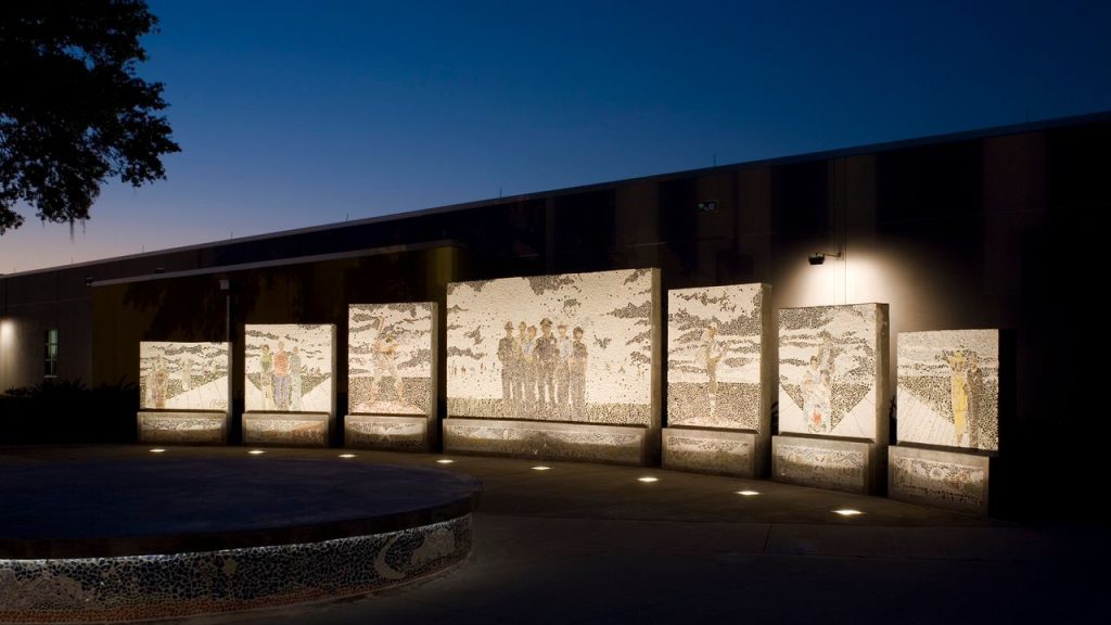 Seven large, illuminated mosaic panels stand outdoors at night against a dark building. Each panel depicts figures and scenes using small, light-colored tesserae against a dark background. A circular, mosaic-covered structure with ground lights is in the foreground.