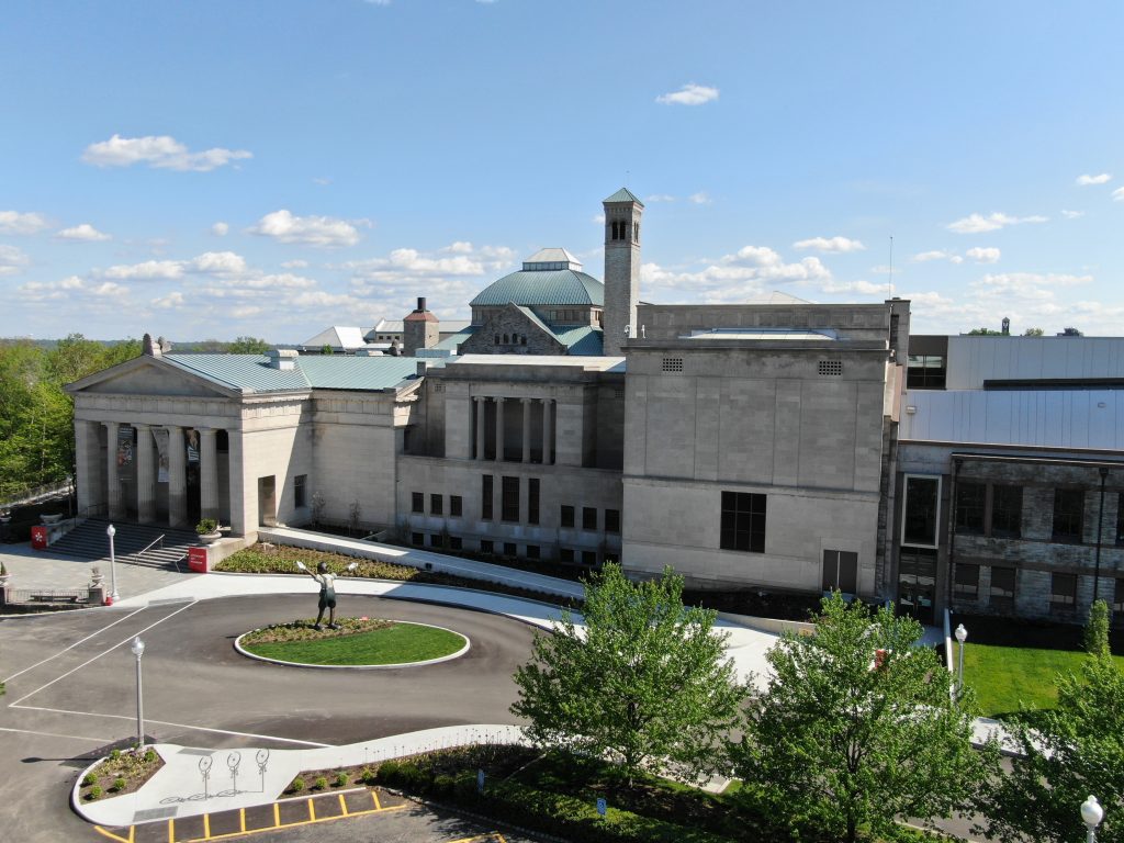 A light-colored stone museum building features a classical facade with columns, a domed roof, and a bell tower. A circular driveway in the foreground includes a statue of a person with outstretched arms holding white objects.