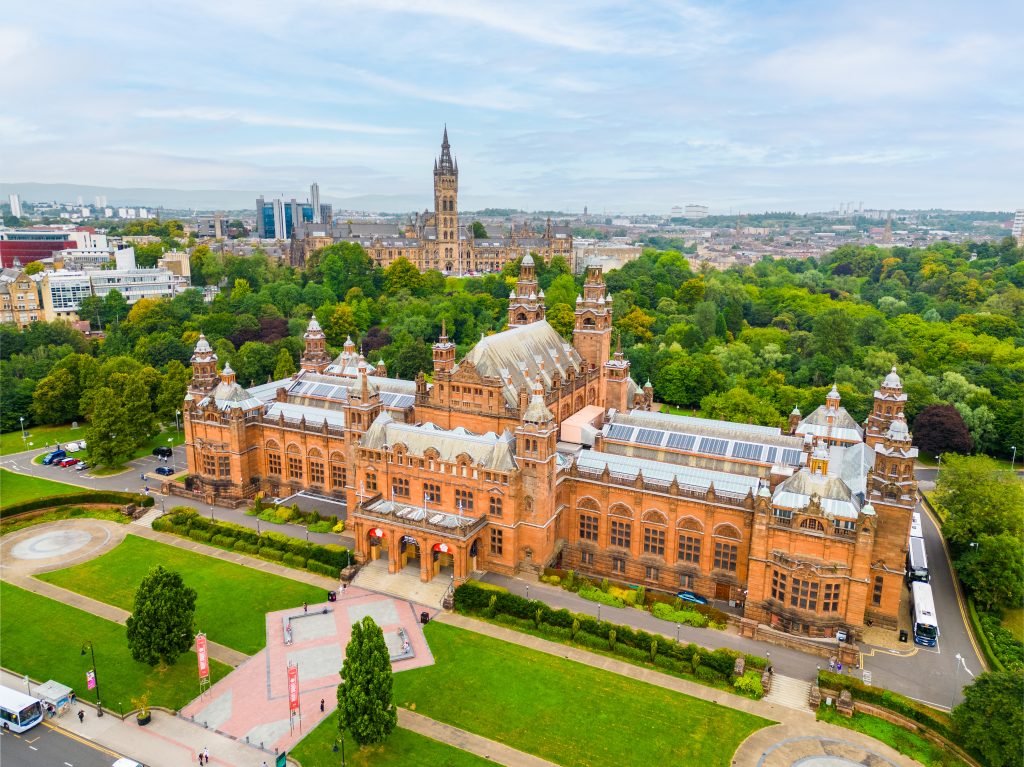 An aerial view shows the large, ornate reddish-brown Kelvingrove Art Gallery and Museum, featuring multiple towers and arched entrances, surrounded by green lawns and trees. In the background, the spired Glasgow University building rises above a cityscape.
