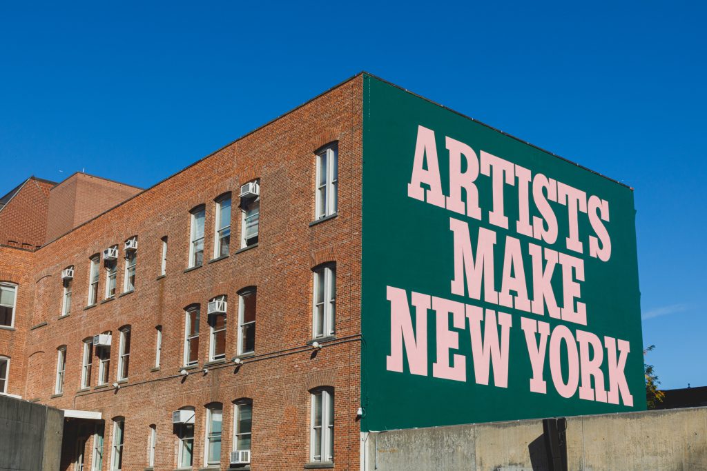A brick building with many windows stands against a clear blue sky. One side of the building is painted green with large pink text that reads "ARTISTS MAKE NEW YORK."