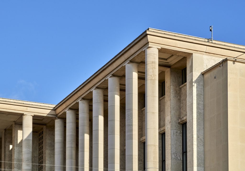 A light-colored stone building with several tall, round columns stands under a clear blue sky, with sunlight casting strong shadows on the columns.