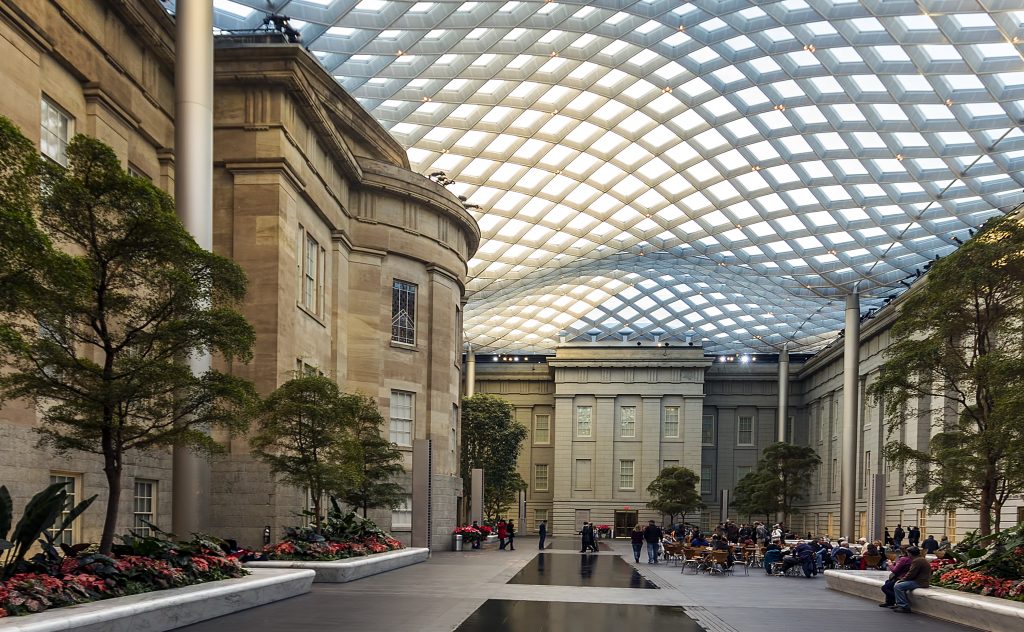 A vast indoor atrium features classical stone buildings and a large, grid-patterned glass ceiling. Trees and flower beds are interspersed with people walking and seated at cafe tables and benches around a reflective floor.