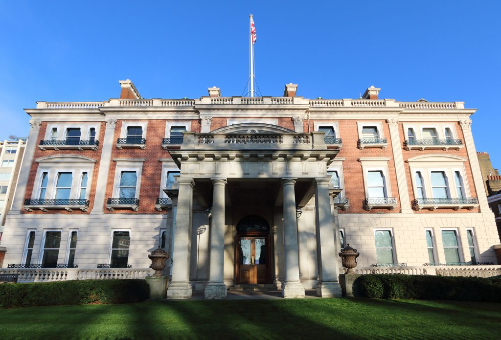 A grand, multi-story building with red brick walls and light-colored stone trim stands under a clear blue sky. It features a prominent entrance portico with columns and a balcony, and a flag flies from the roof. A manicured green lawn and hedges are in the foreground.