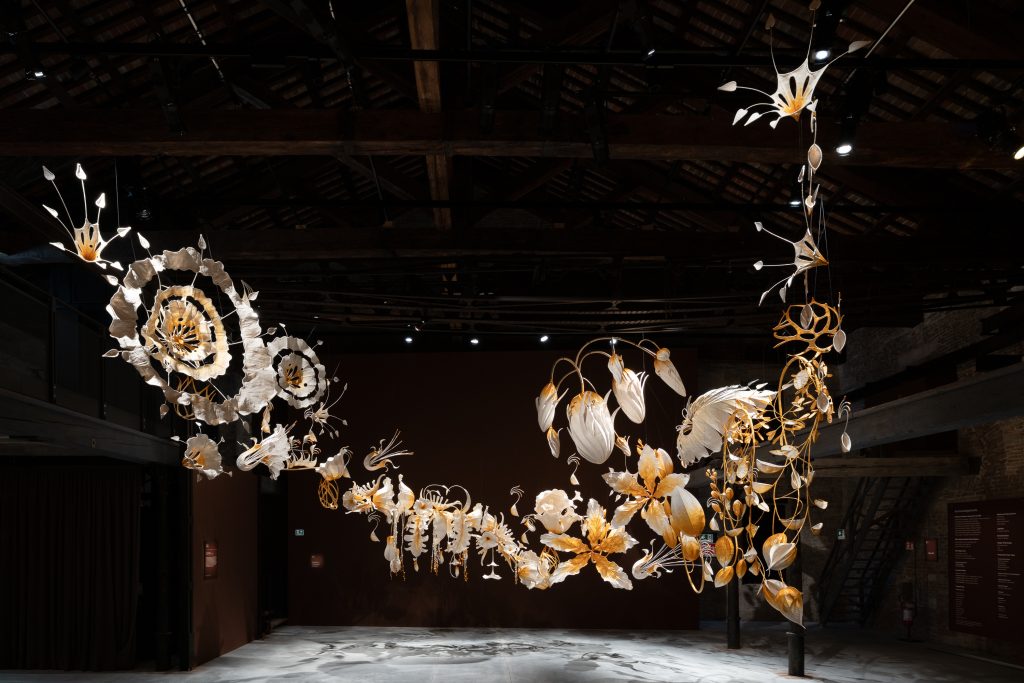 A large, intricate sculpture made of numerous white and golden organic shapes hangs from a dark, beamed ceiling. The sculpture forms a wide, curved arc, resembling a flowing garland of abstract leaves and flowers.