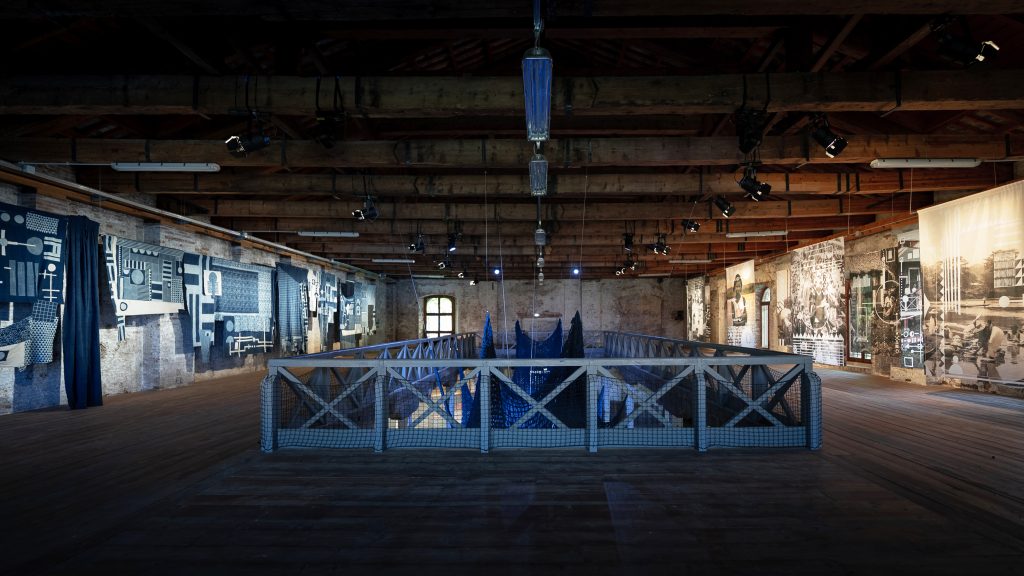 A dimly lit exhibition space with wooden floors and exposed brick walls. In the center, a raised platform with white railings encloses a dark blue net sculpture. Walls are adorned with fabric panels featuring blue and white geometric patterns and large banners with black and white photographs.