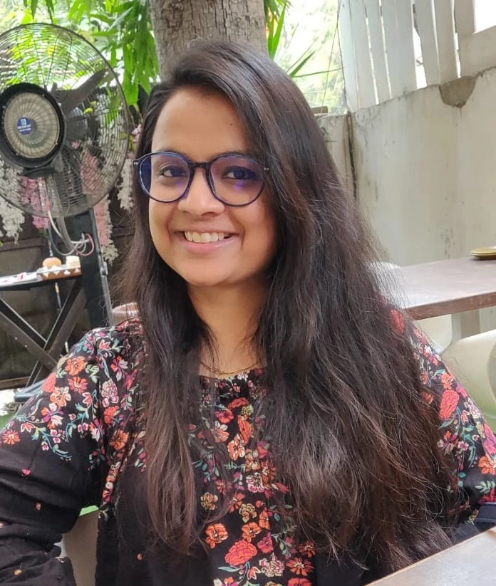 Ankita Tapadia sitting outside in front of a tree, with a fan in the background.
