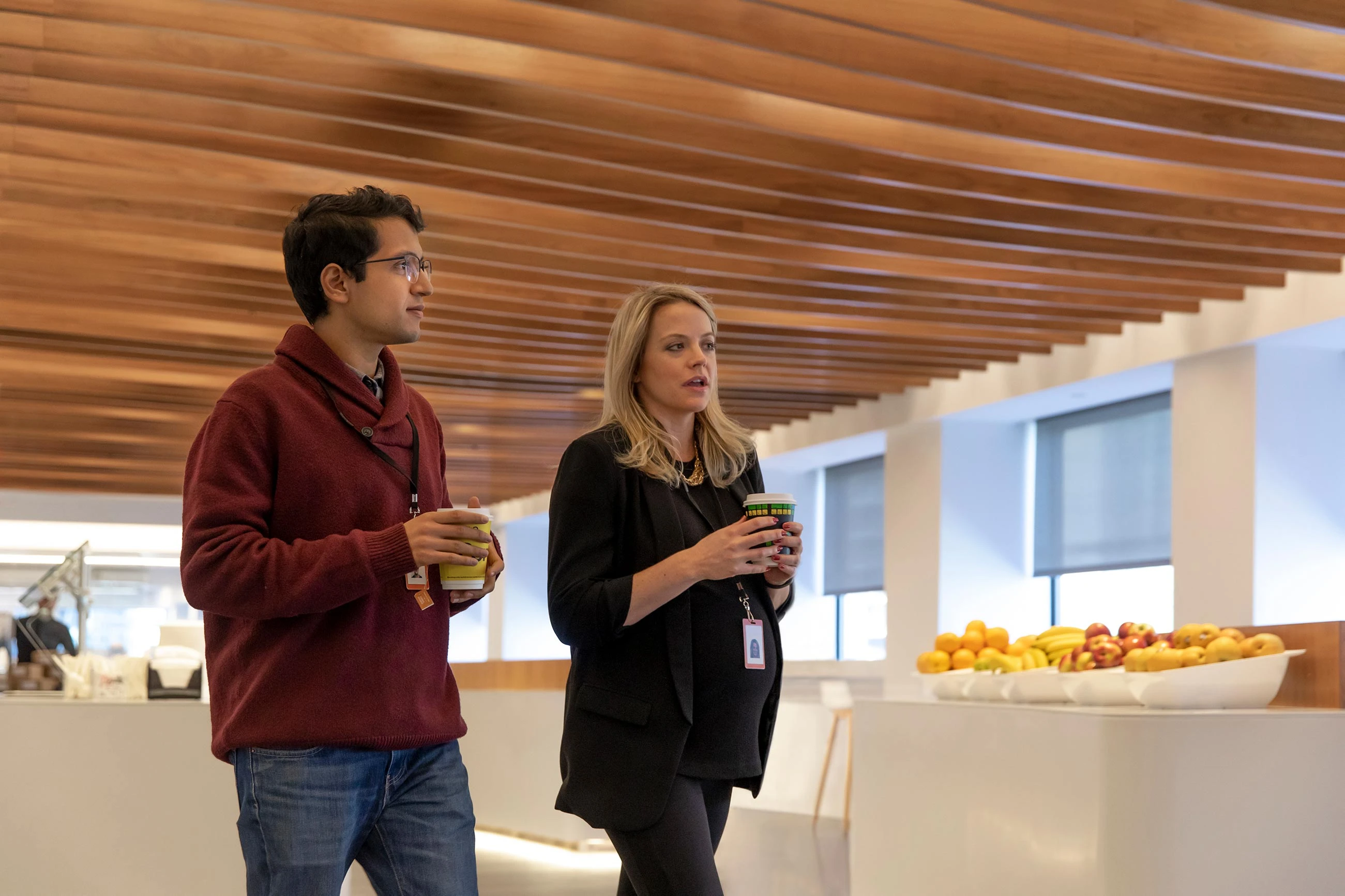 Two Bloomberg employees, one of them pregnant, discuss a project while walking through the office with warm drinks.