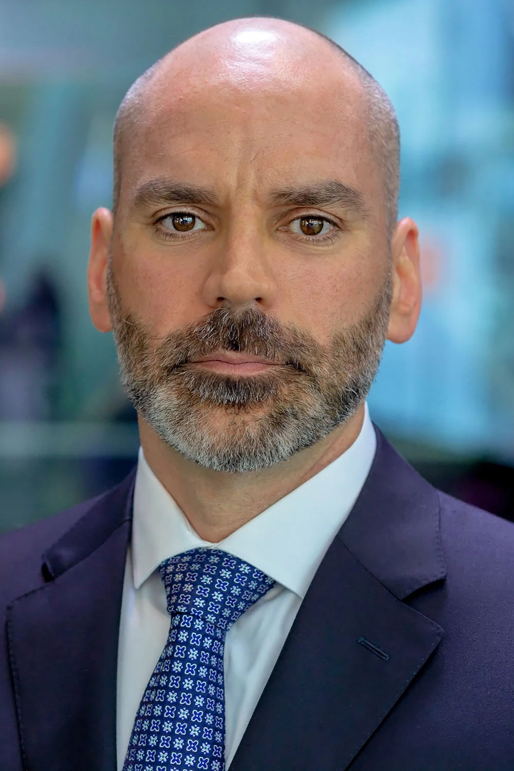 Shawn Edwards poses for a headshot. He is a white male with extremely short hair and a bear, wearing a navy suit with a blue shirt and tie.