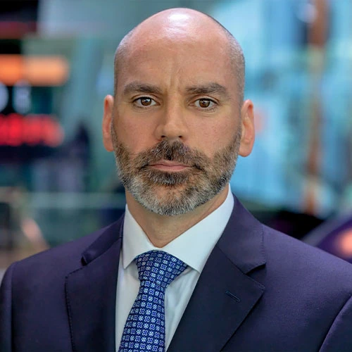 Shawn Edwards poses for a headshot. He is a white male with extremely short hair and a bear, wearing a navy suit with a blue shirt and tie.