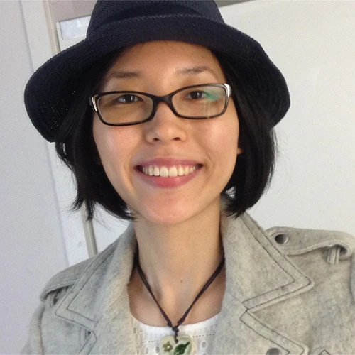 Aram Chung smiles at the camera. She has a dark hat, short hair, and is wearing an off-white denim jacket with a white top and necklace.