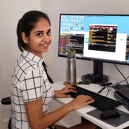 Bhavya Kumari stands at her workstation. She has her long, dark hair pulled back in a ponytail and is wearing a white collared shirt with a large black windowpane pattern.