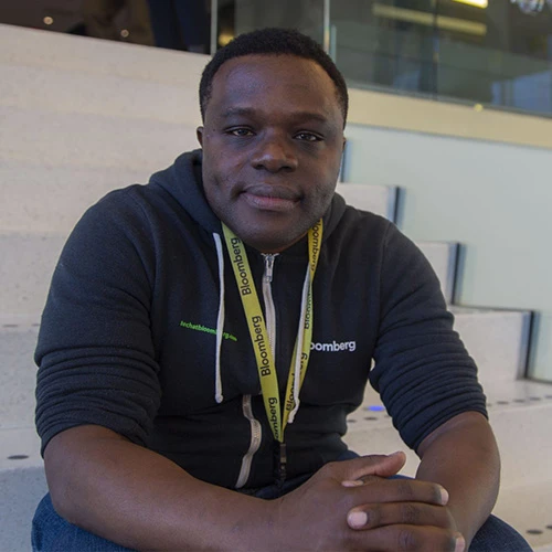 Deji Akinyemi sits in a stadium seat. He is clean-shaven with short-cropped dark hair and is wearing a dark-gray zip-up hoodie with a white Bloomberg logo on the left chest.