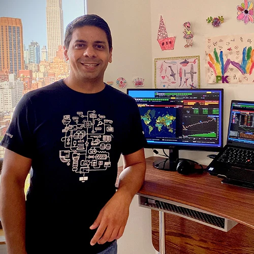 Pratik Karia leans on his work-from-home desk and smiles at the camera. He's wearing an art-laden black tee shirt and is clean-shaven with short, dark hair.