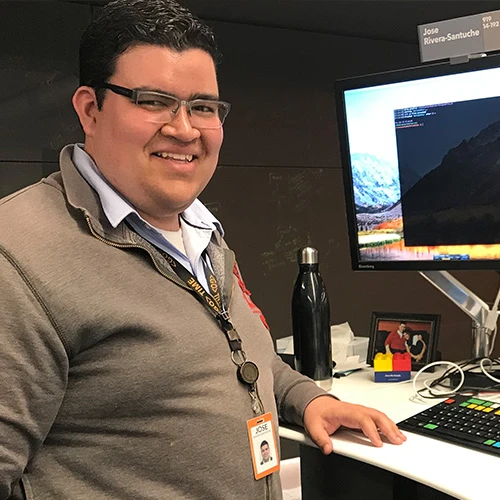 José Rivera-Santuche stands in front of his workstation at Bloomberg. He has finely-styled black hair and rimless glasses and is wearing an olive quarter-zip sweater on top of a light-blue collared shirt.