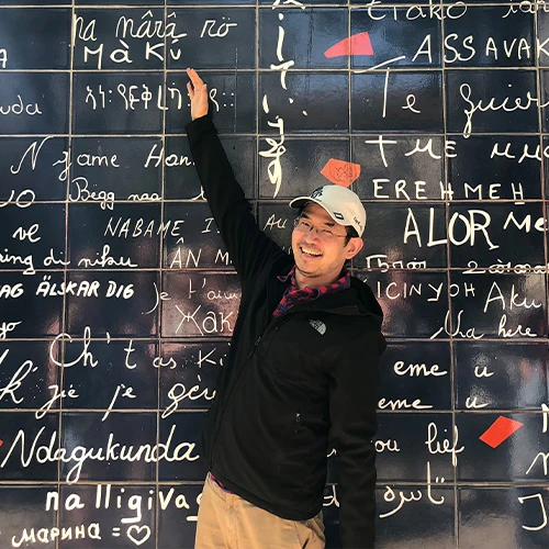 Conway Tang stands in front of a wall, pointing to a piece of white hand-written text on top of the dark chalkboard-like wall. He wears a white baseball hat, a black The North Face jacket, and khaki chinos.