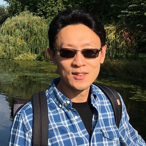 Keyong Li stands in front of a calm pond. He's wearing a backpack on top of a light blue plaid button-up shirt and dark transition lenses.