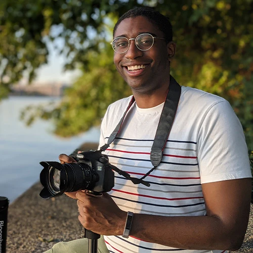 Meshach Jones smiles at the camera and holds his personal DSLR camera ready for his next short. He is clean-shaven with short, black hair and is wearing a white tee shirt with multi-colored horizontal stripes across the body.