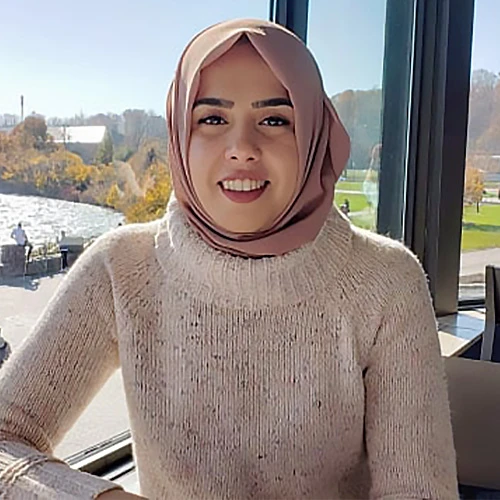 Tuba Opel stands in front of a large glass window on a high floor. She smiles at the camera and is wearing a white sweater and a light-pink hijab.