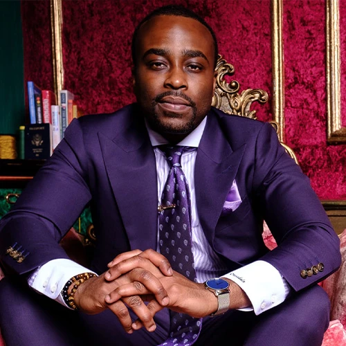 Terrence Crossdale sits in a plush pink velvet throne. He's wearing a stunning, athletic-cut lapis suit with white cuffs, and a purple tie and pocket silk. He has a well-groomed beard and short-cropped hair.
