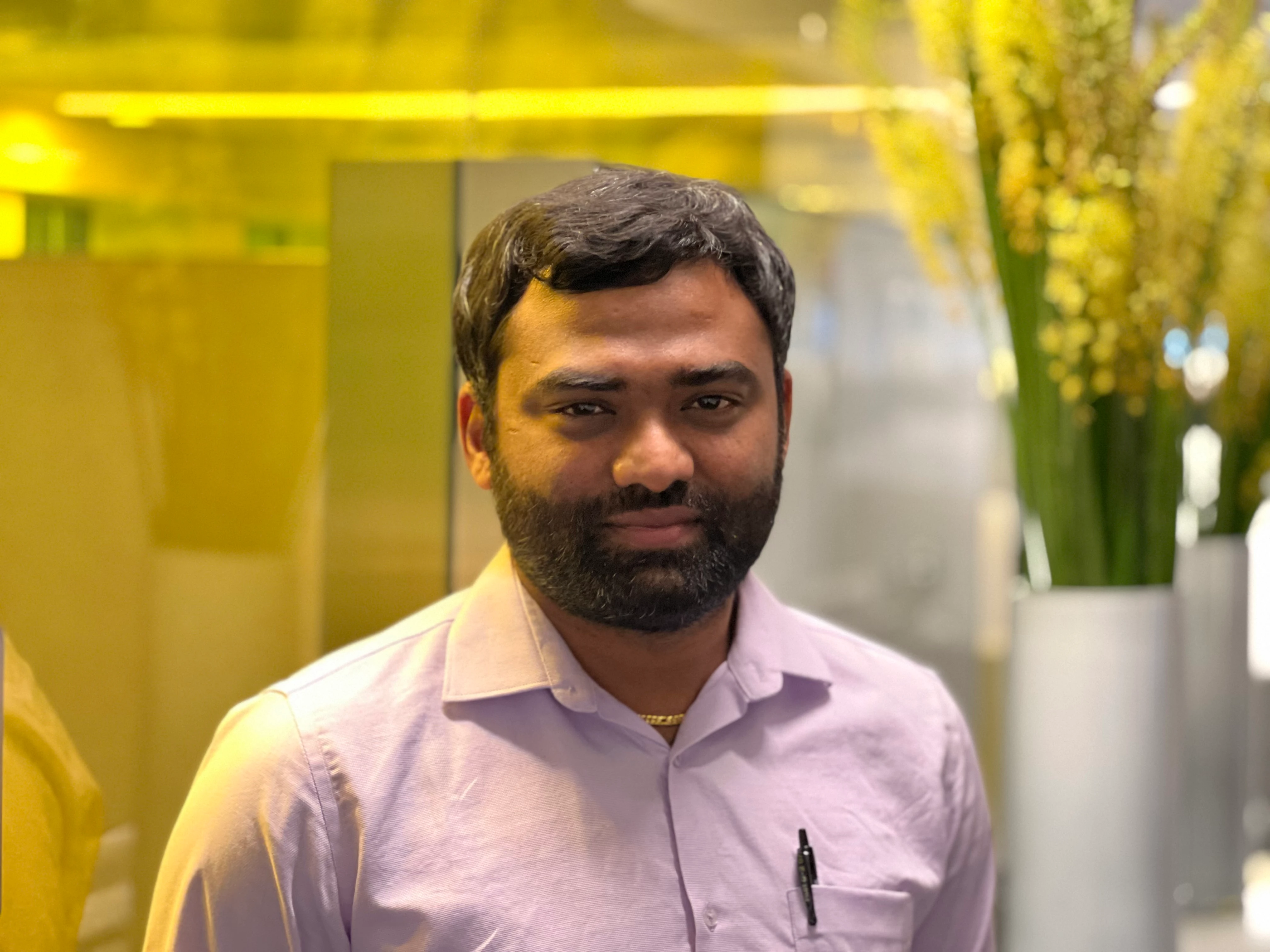 Gopi Sivashanmugam standing in front of a yellow glass wall and an arrangement of yellow flowers in the Bloomberg office in NYC