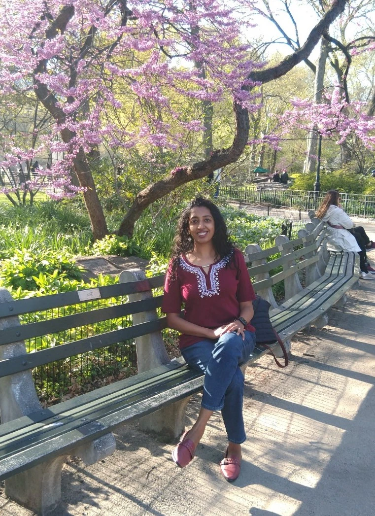 Shreya Krishnan sitting on a park bench in front of a blooming tree.