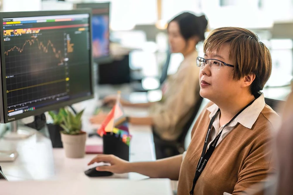 What-we-do A woman at the Bloomberg Singapore office works on the Bloomberg Terminal as a part of her job. She's wearing a tan sweater with a beige blouse, dark-rimmed glasses, ear-length light brown hair, and a Bloomberg lanyard with her employee ID.