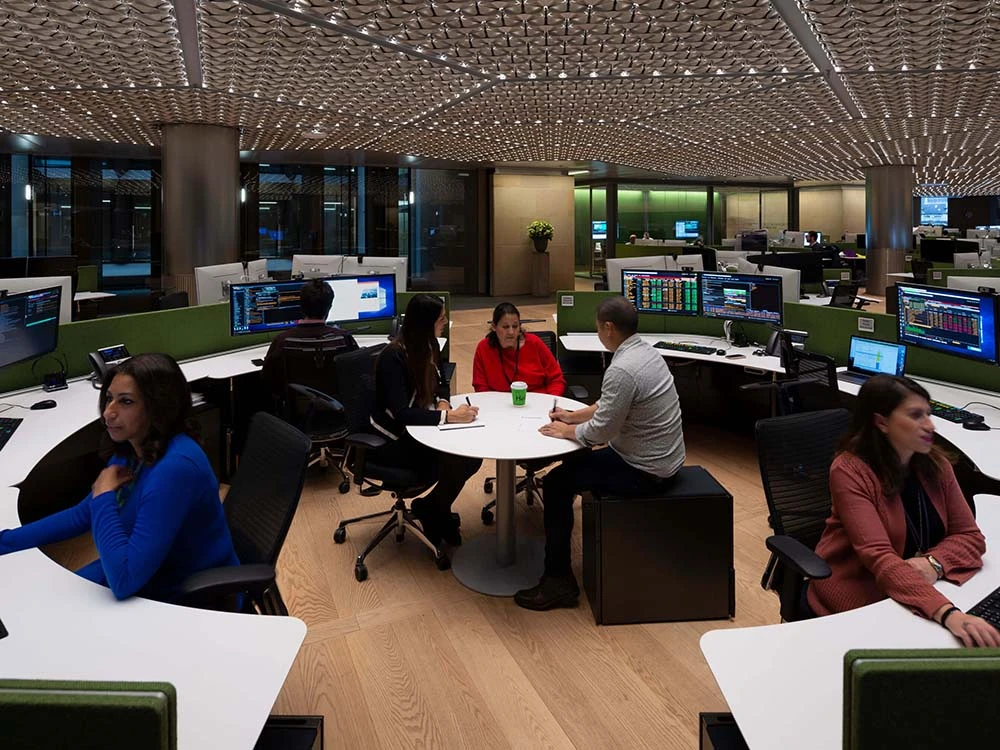 Bloomberg-London-office-collaboration-desk-pods A group of Bloomberg London office employees sync at a center round table as a series of desks wrap around them.
