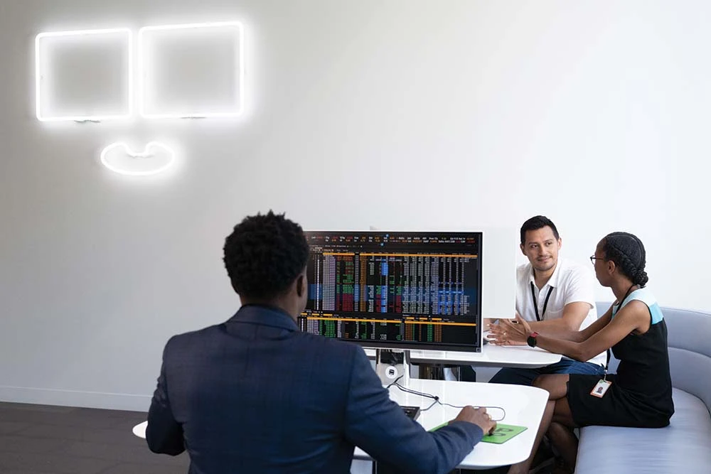 What-we-do-at-Bloombergs-New-York-offices A white neon sign at Bloomberg's 731 Lexington headquarters building glows in the background as a Black employee works on a Bloomberg Terminal in the foreground while a male and female employee collaborate in the background.